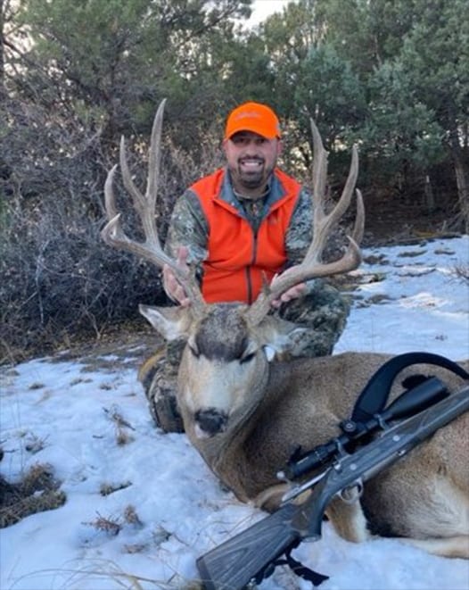 A hunter in an orange vest poses with a harvested mule deer in a snowy, wooded area, with a rifle lying next to the deer.