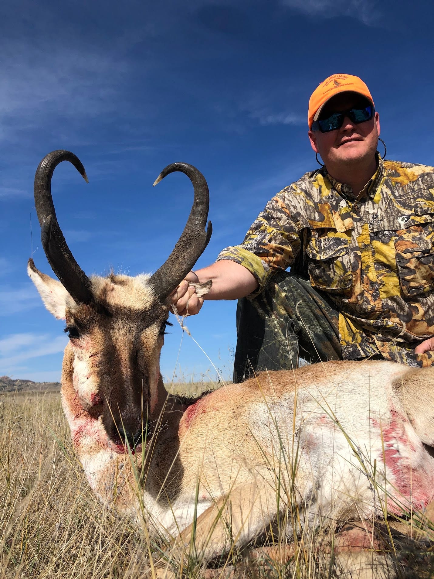 A hunter poses with a harvested pronghorn in a grassy field under a blue sky.