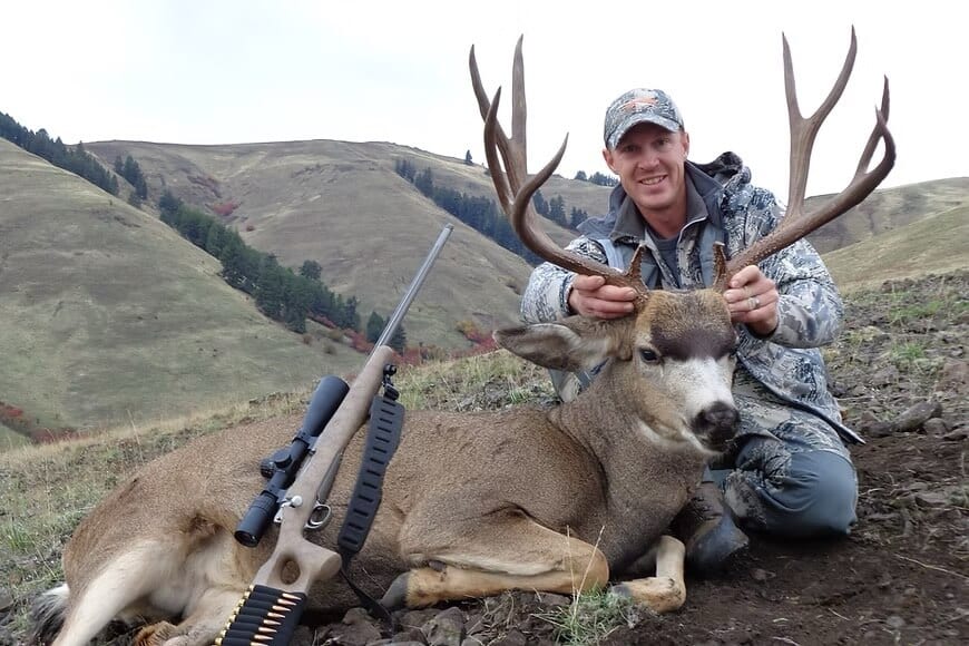 A hunter poses with a harvested mule deer and a rifle in a hilly, open landscape.