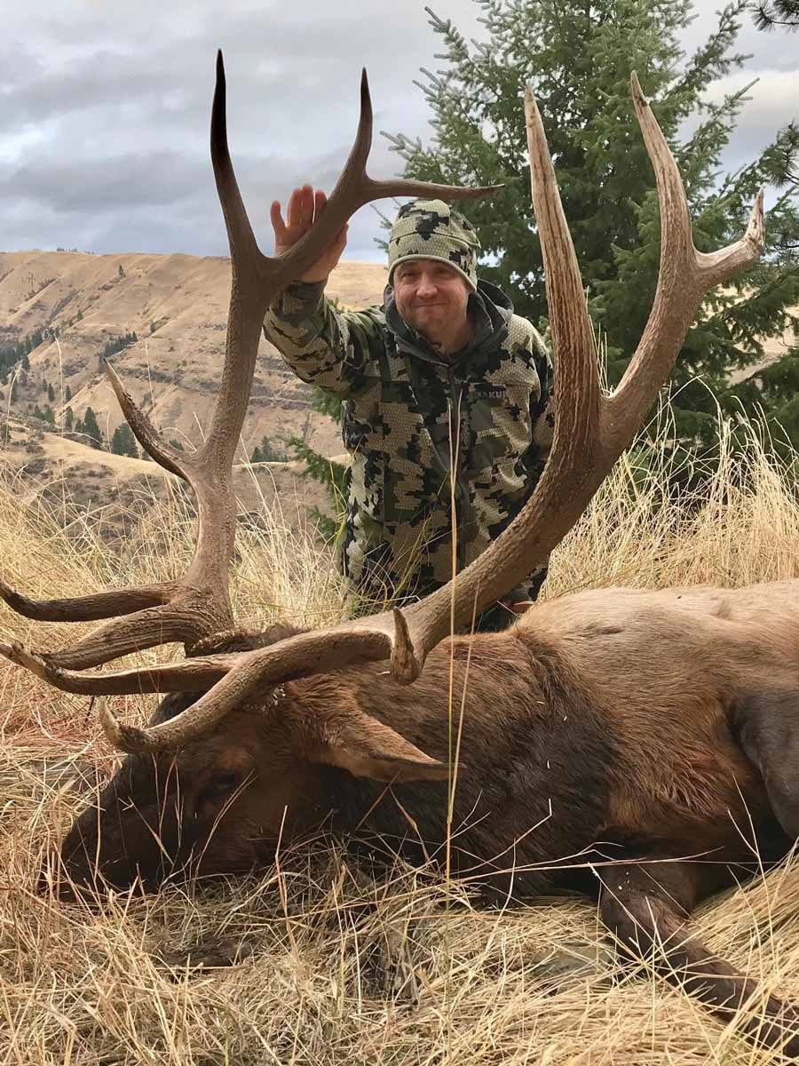 A hunter in camouflage poses with a large harvested elk in a grassy field.