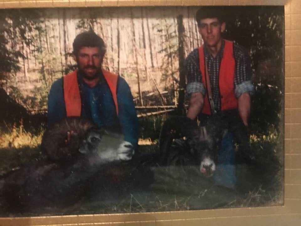 Two hunters pose with two harvested bighorn sheep in a wooded area.