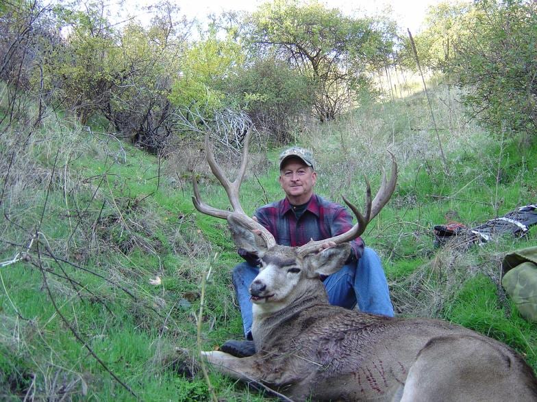 A hunter in a camouflage hat and plaid shirt poses with a harvested mule deer in a grassy, brushy area.