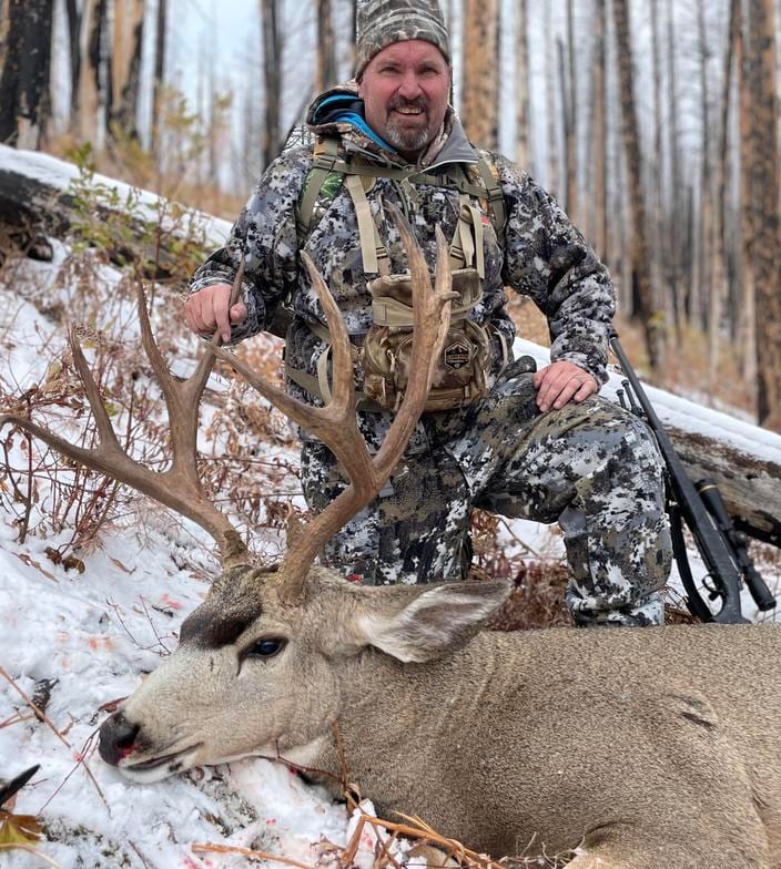 A hunter kneels in the snow next to a harvested mule deer in a forest.