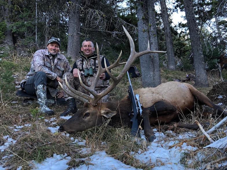 Two hunters pose with a harvested elk and a rifle in a snowy, wooded area.