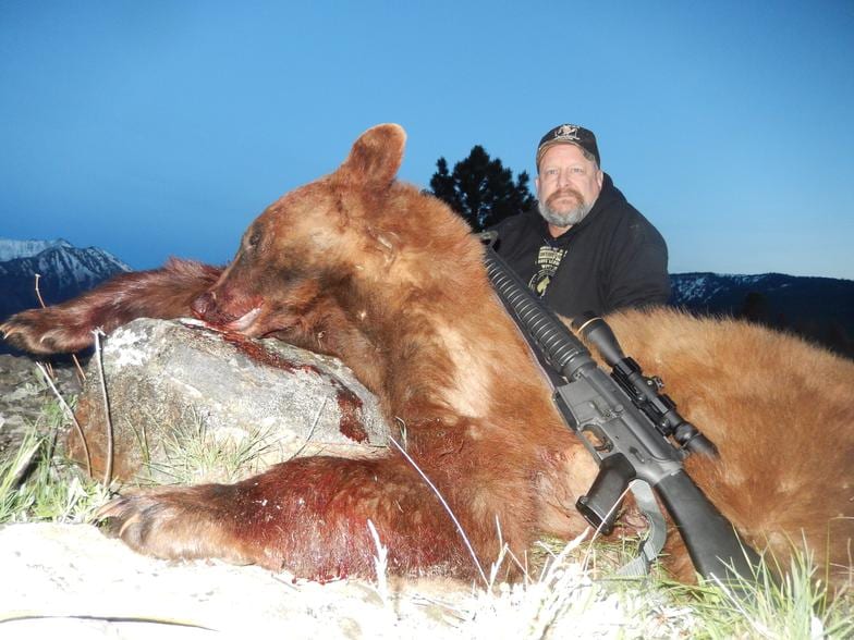 A hunter poses with a harvested black bear and a rifle against a mountain backdrop.