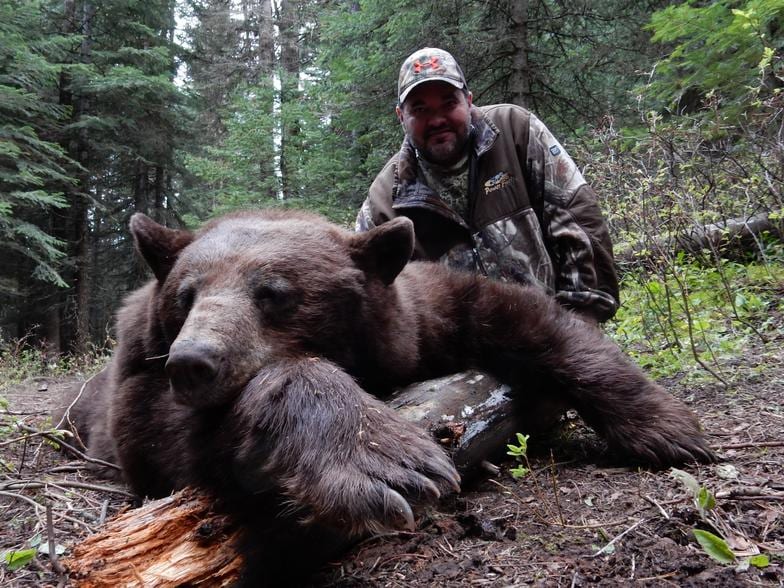 A hunter poses with a harvested bear in a forest.