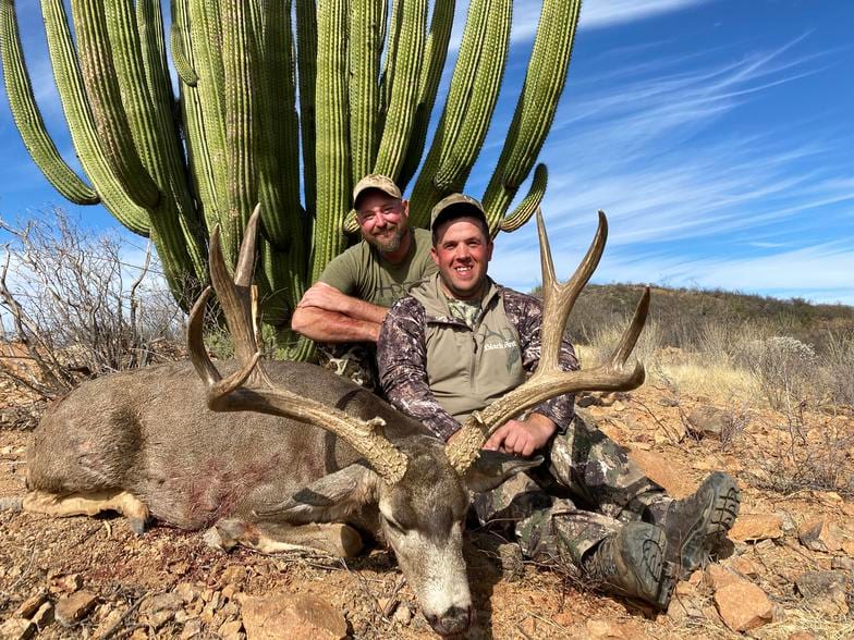 Two hunters pose with a harvested mule deer in front of a large saguaro cactus.