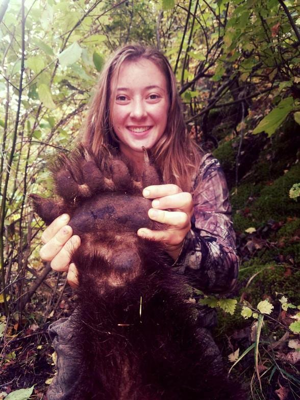 A woman in camouflage smiles while holding a black bear paw in a wooded area.