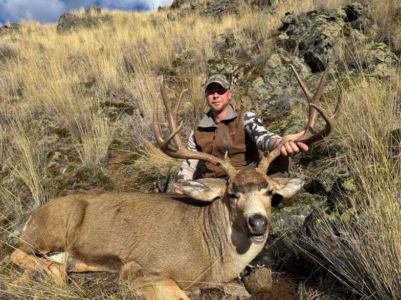 A hunter poses with a harvested mule deer in a grassy, rocky landscape.