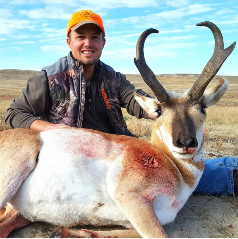 A hunter in a camouflage vest and orange hat poses with a harvested pronghorn in a field.