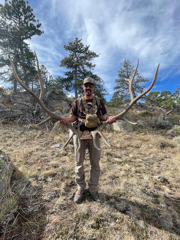 A man holds a large set of elk antlers in a natural, outdoor setting.