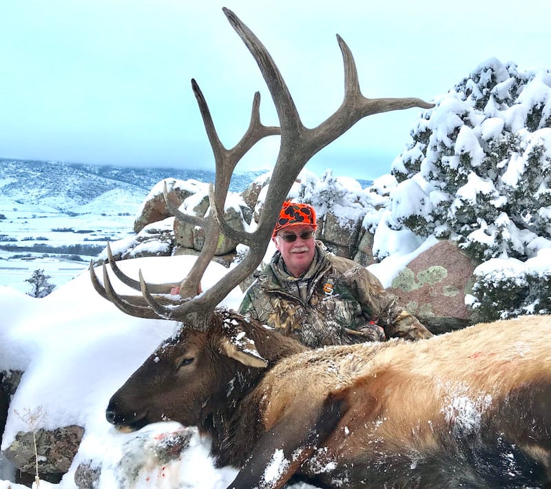 A hunter poses with a harvested elk in a snowy, mountainous landscape.