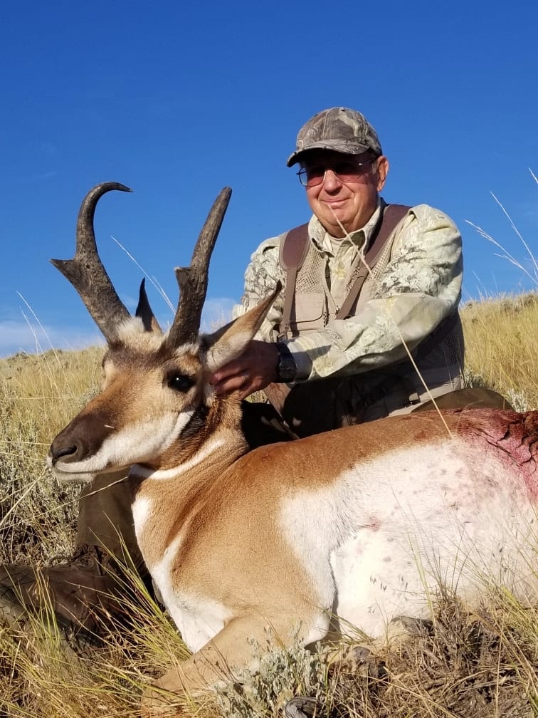 A hunter in camouflage poses with a harvested pronghorn in a grassy field under a clear blue sky.