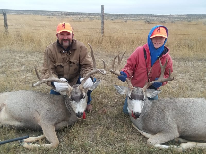 An adult male and a child pose with two harvested mule deer in a field.