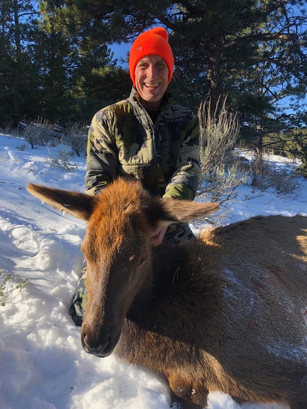 A hunter in a camouflage jacket and orange hat poses with a harvested elk in a snowy, forested landscape.