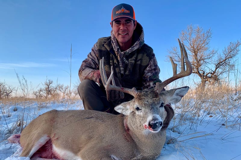 A hunter kneels in the snow next to a harvested mule deer.
