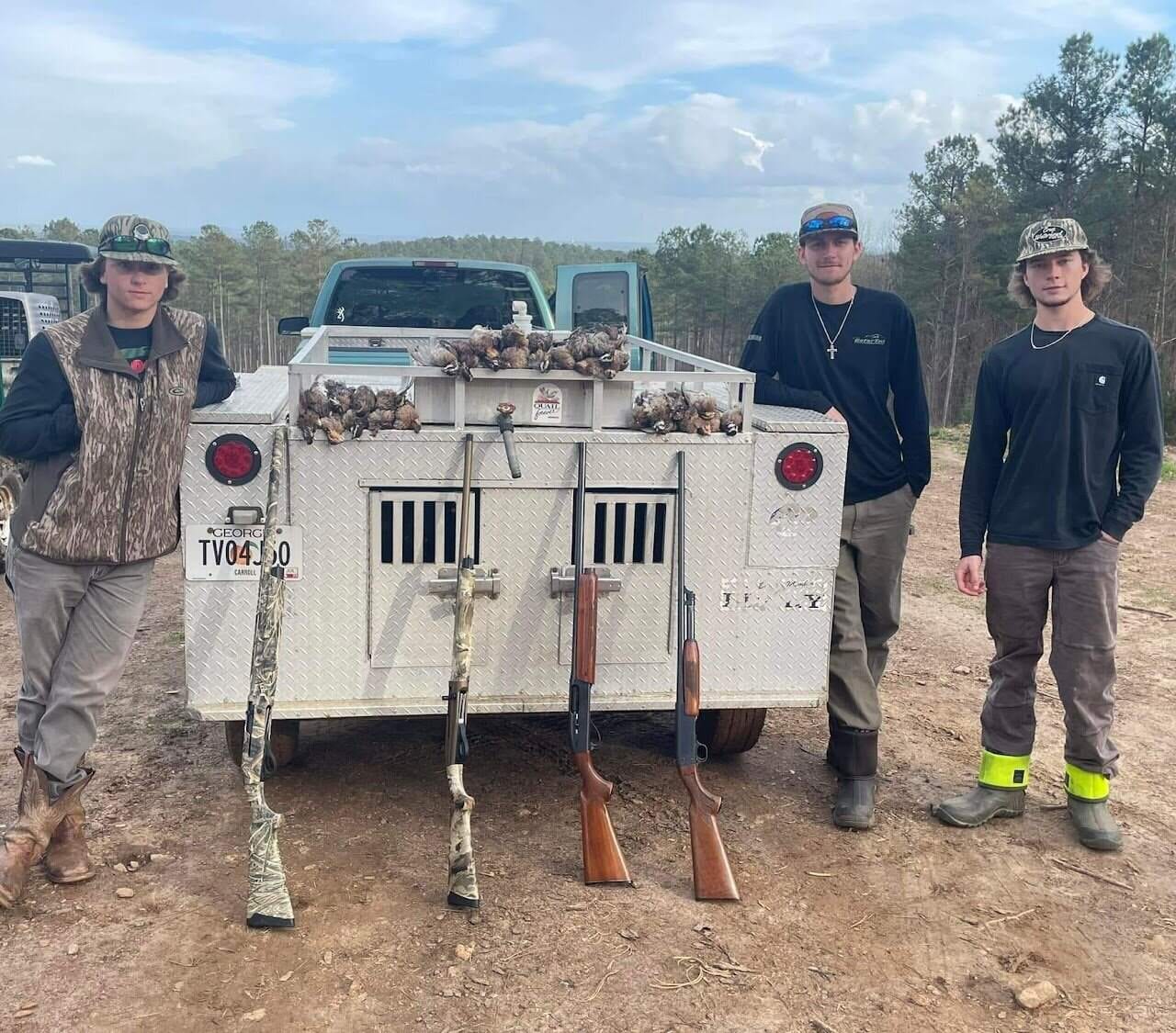 Three young hunters beside an aluminum dog-box trailer with quail and shotguns