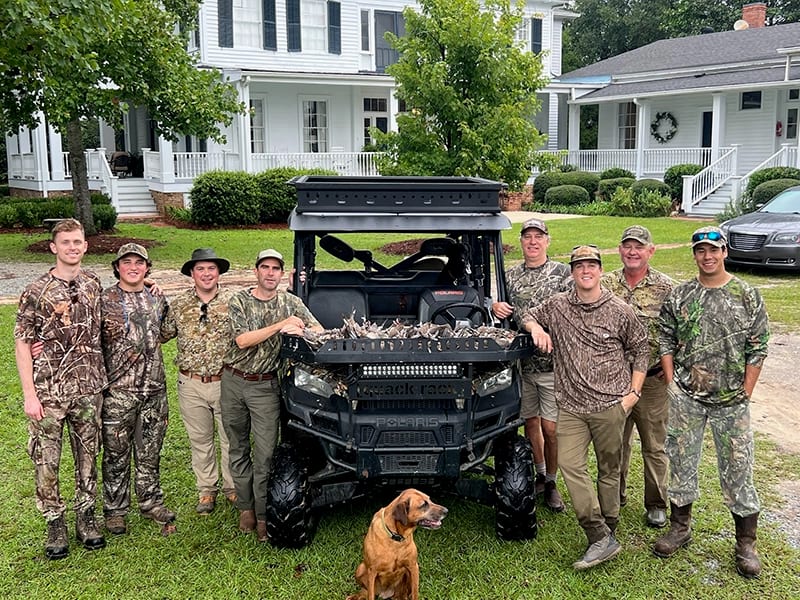 A group of hunters and a dog pose with a UTV loaded with harvested birds in front of a white house.