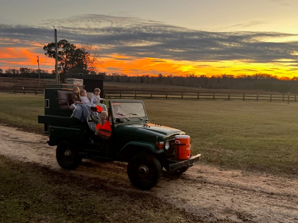 People in a green off-road vehicle on a dirt path at sunset in a field.