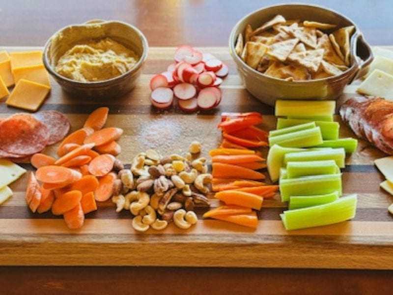 A wooden cutting board is laden with various snacks including cheese, crackers, vegetables, nuts, and dips.
