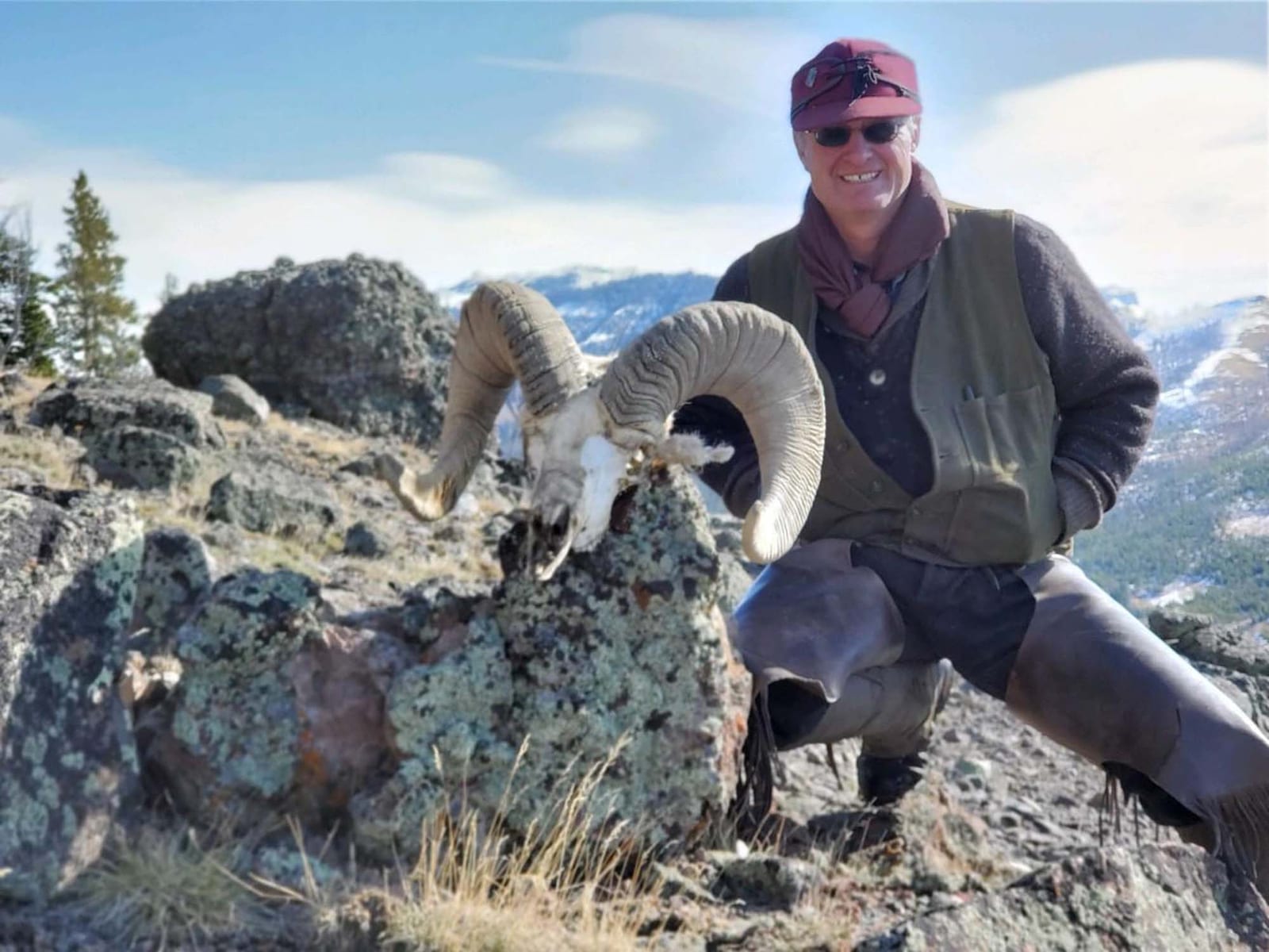 A man poses with a bighorn sheep skull and horns on a rocky mountain.