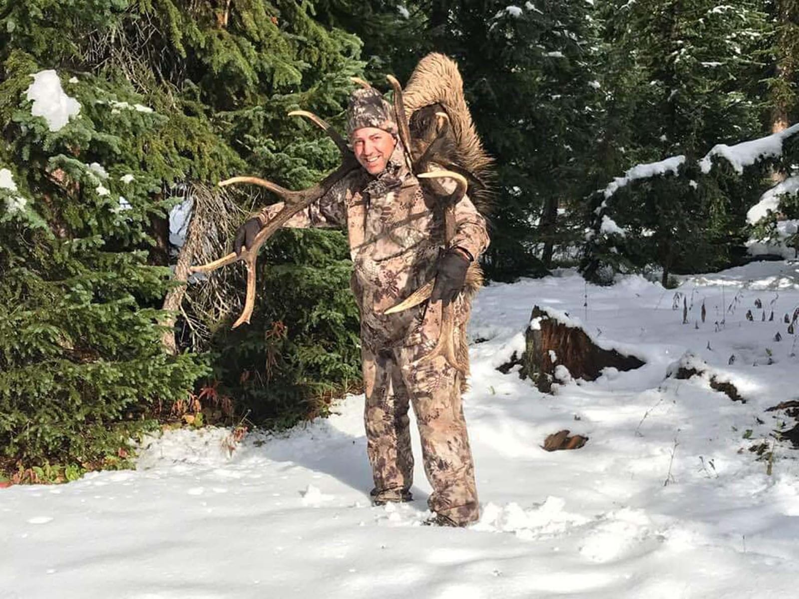 A hunter in camouflage poses with harvested elk antlers and a bighorn sheep in a snowy forest.