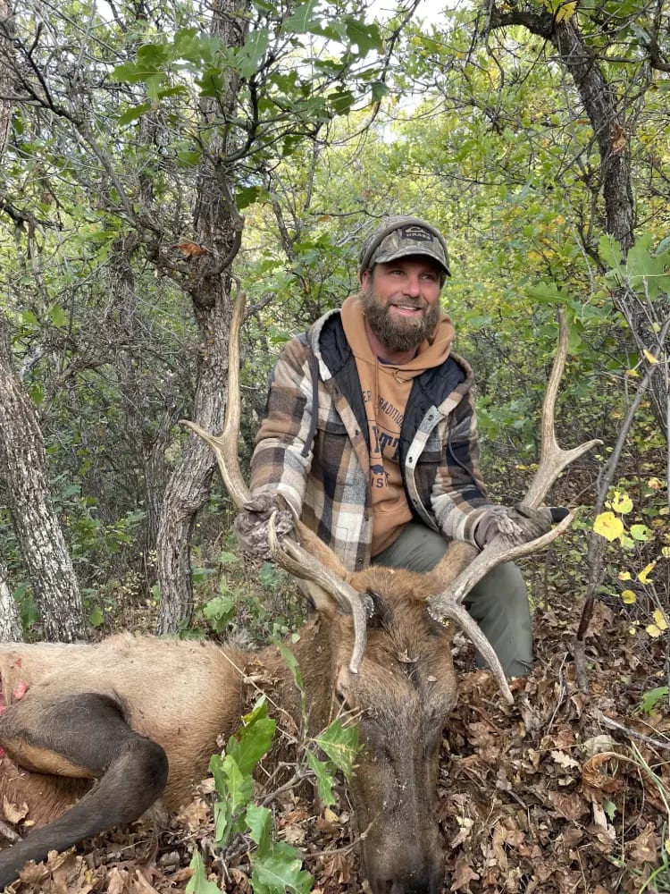 A hunter with a beard and a plaid jacket poses with a harvested elk in a wooded area.