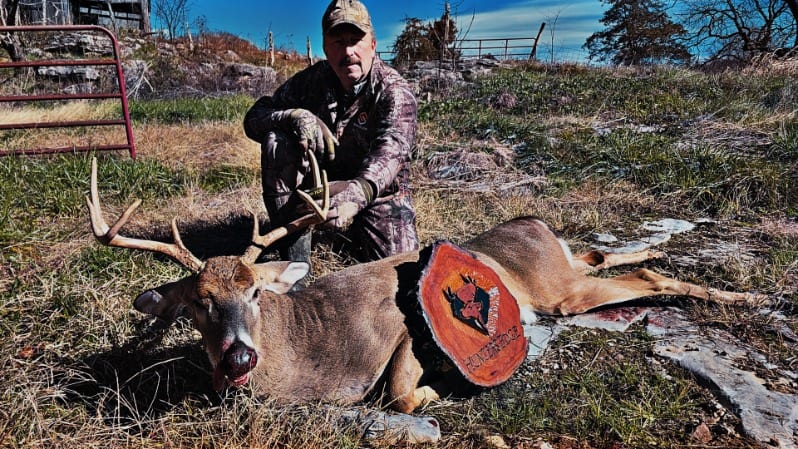 A hunter kneels beside a harvested whitetail deer in a grassy field.