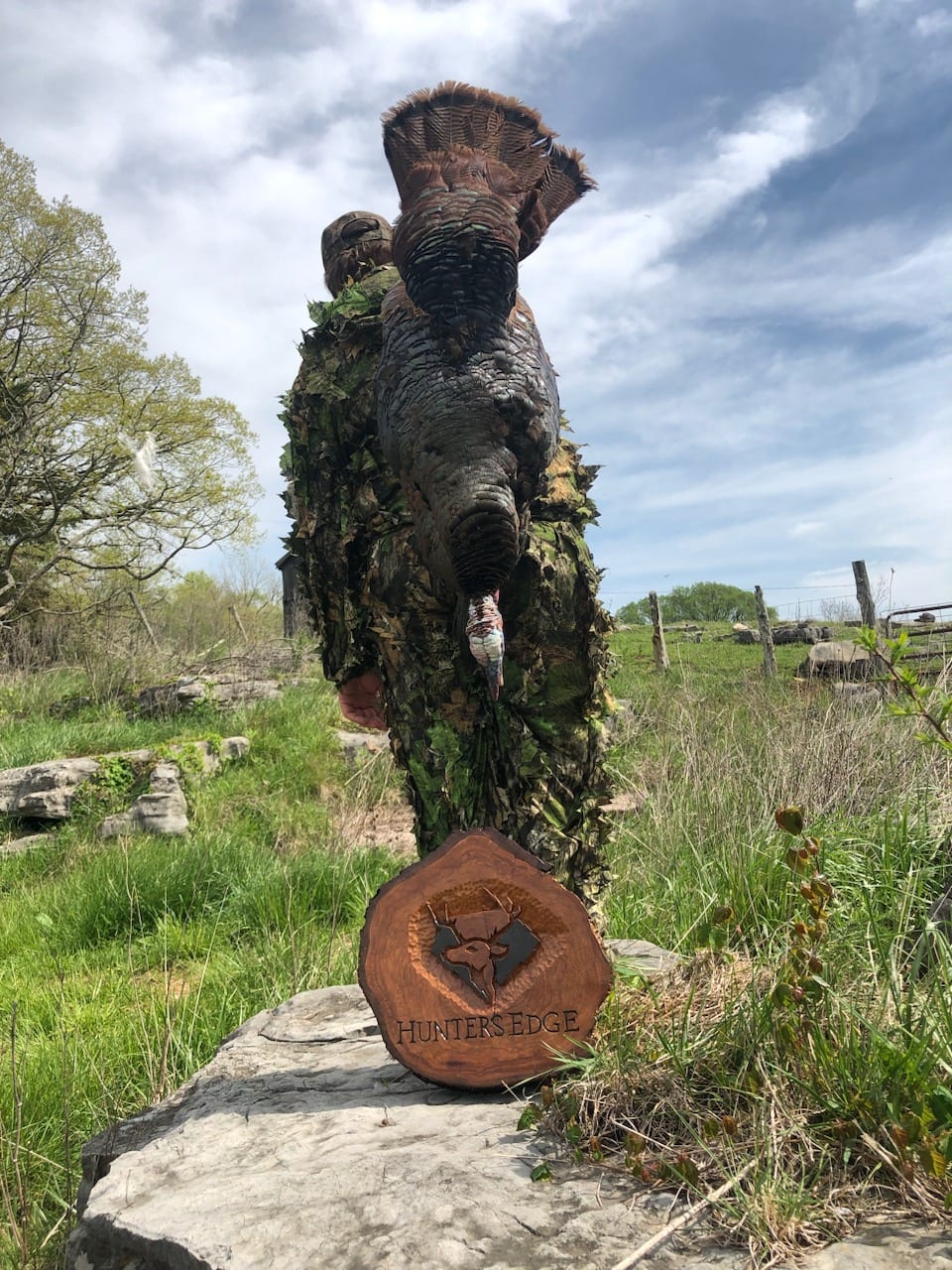 A hunter in camouflage holds a harvested wild turkey in a field, with a 'Hunters Edge' plaque on a rock in the foreground.