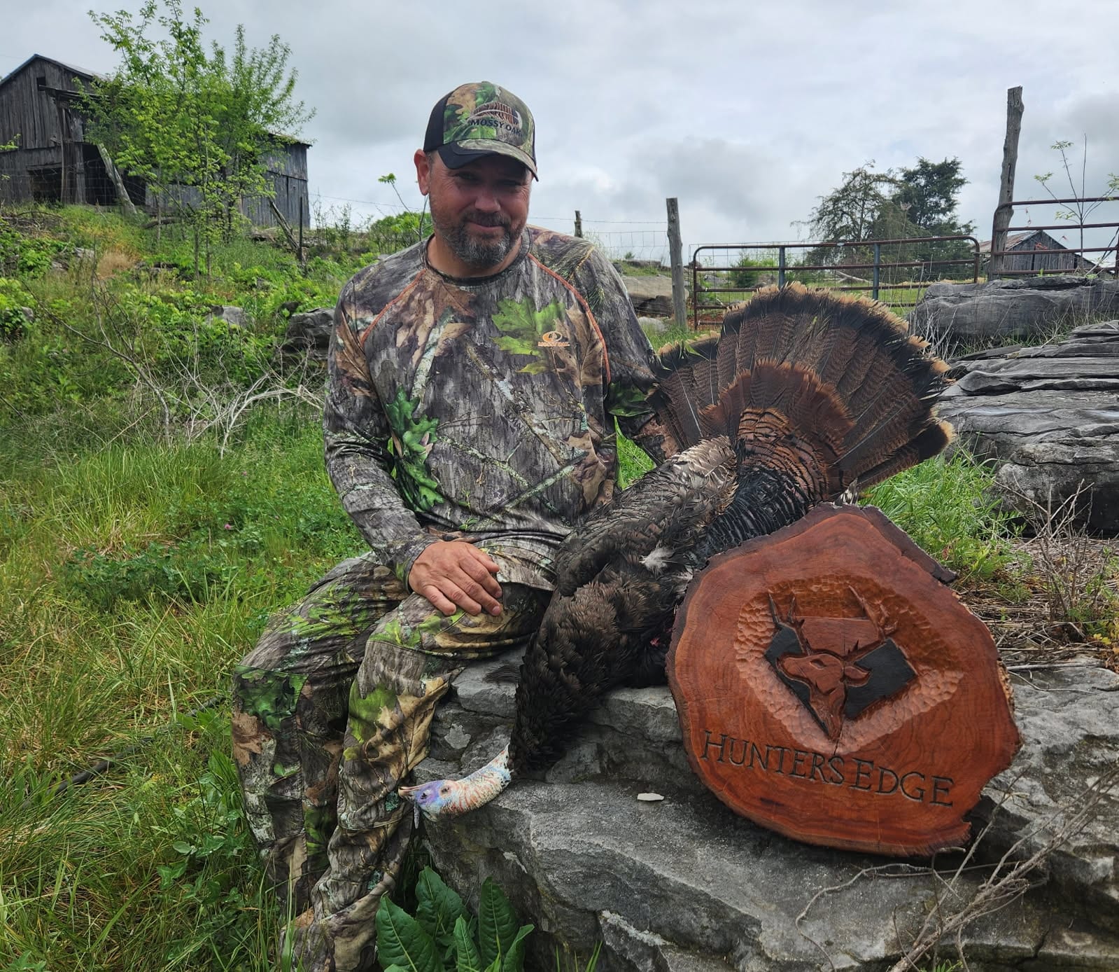 A man in camouflage sits on rocks next to a harvested wild turkey and a wooden plaque.