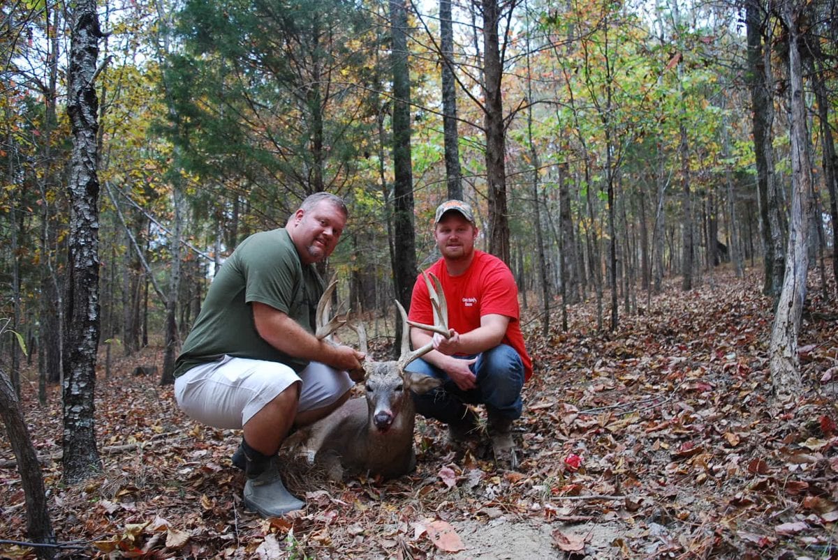 Two hunters pose with a harvested whitetail deer in a forest.
