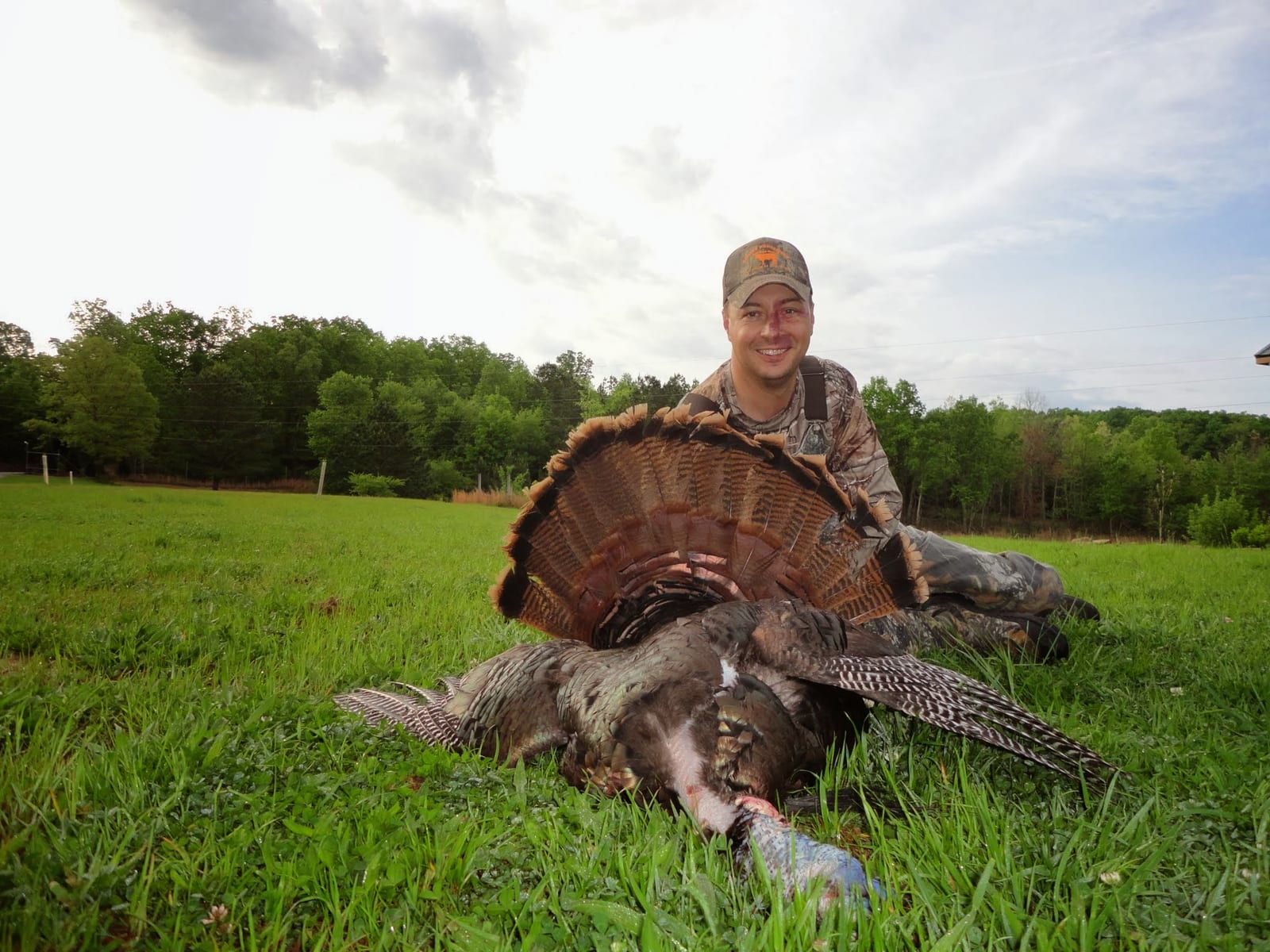 A hunter poses with a harvested wild turkey in a grassy field.