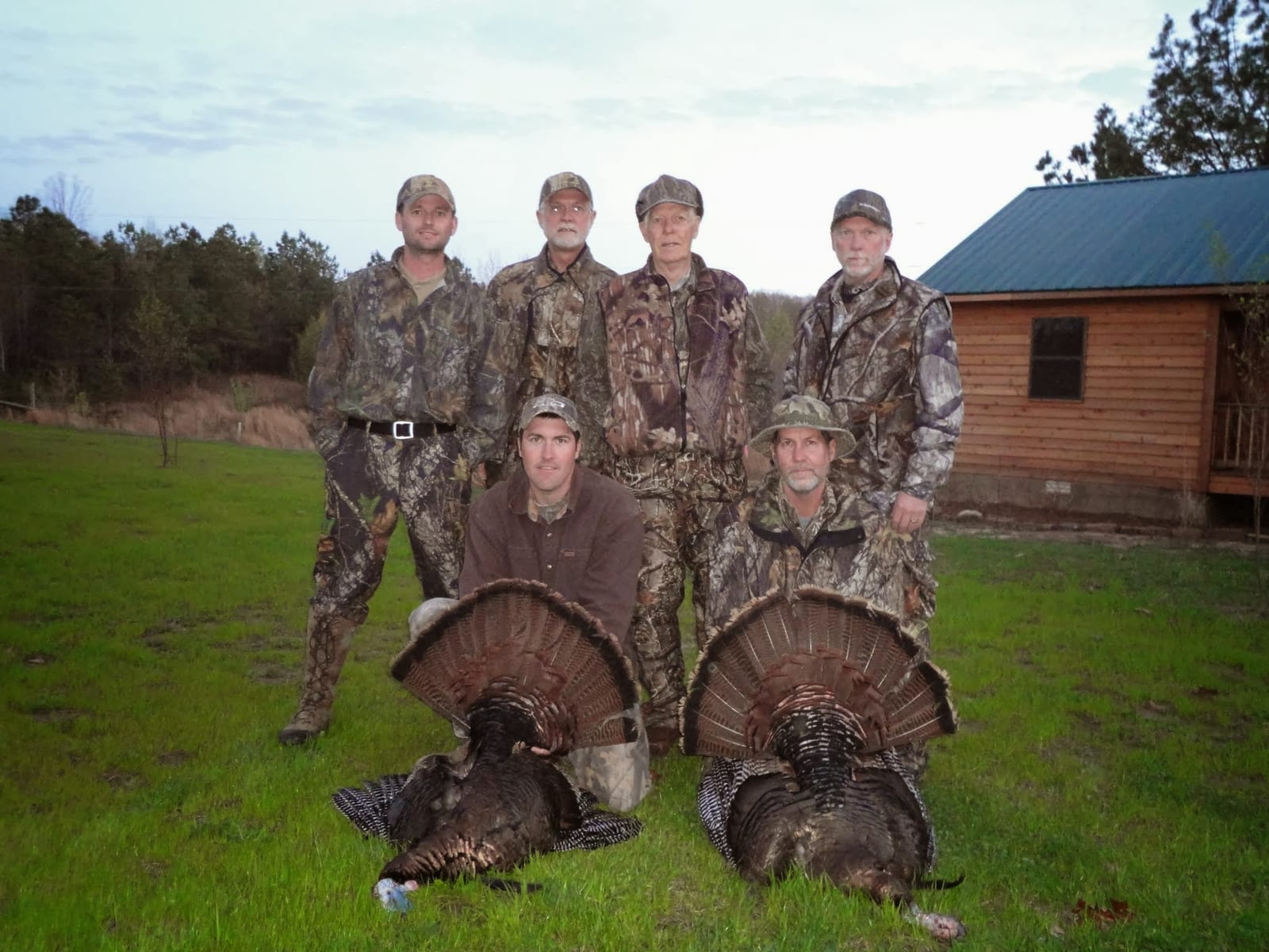 Several hunters pose with two harvested wild turkeys in a grassy field in front of a cabin.