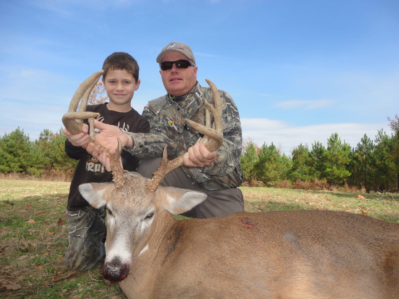 A man and a boy pose with a harvested whitetail deer in a field.