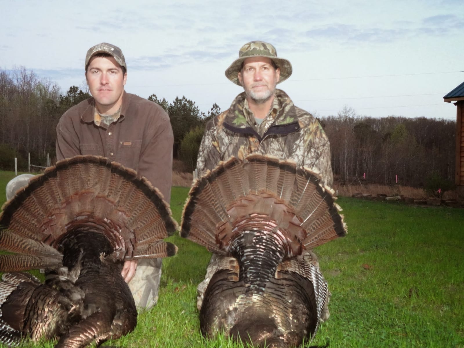 Two hunters pose with their harvested wild turkeys in a grassy field.