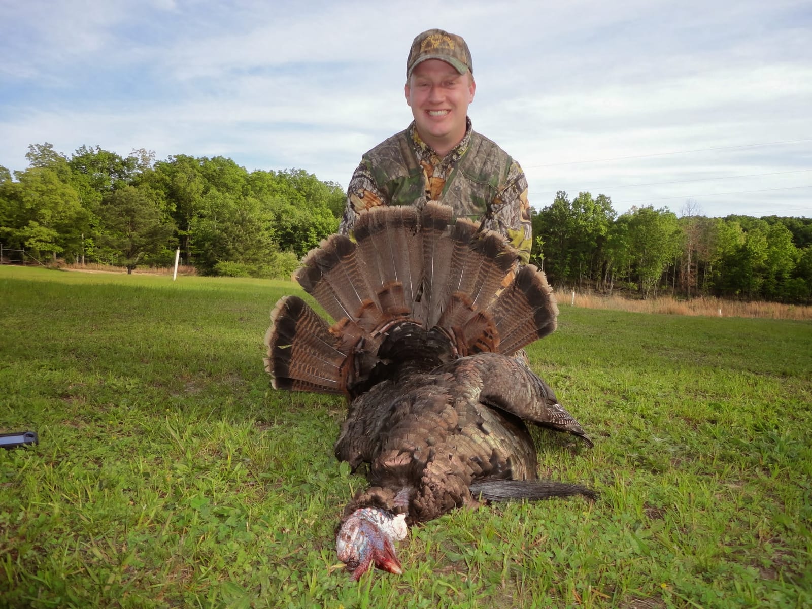 A man in camouflage poses with a harvested wild turkey in a grassy field.