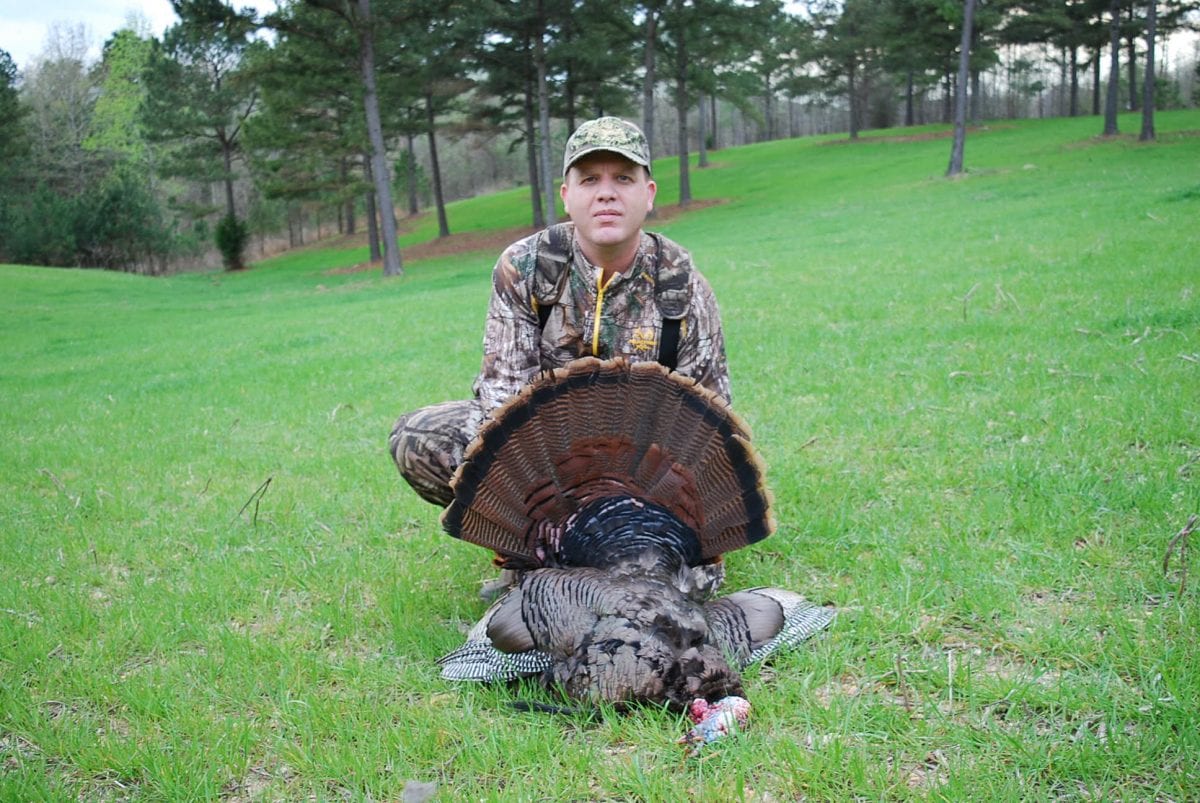 A hunter in camouflage poses with a harvested wild turkey in a grassy field.