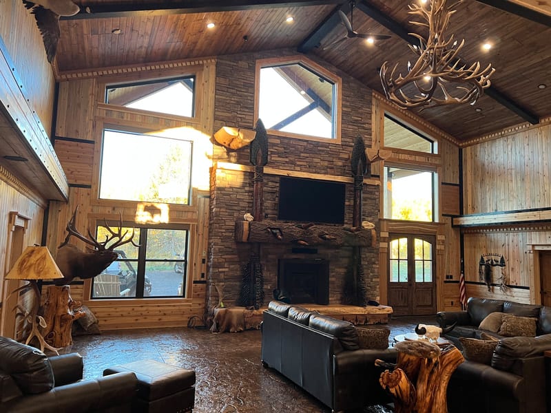 The interior of a rustic lodge with a large stone fireplace, high ceilings, and an elk taxidermy mount.