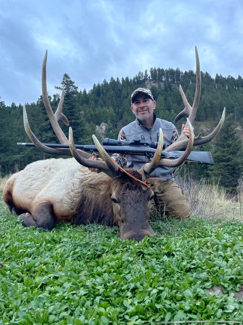 A hunter kneels behind a harvested elk with large antlers, holding a rifle, in a grassy field with trees in the background.