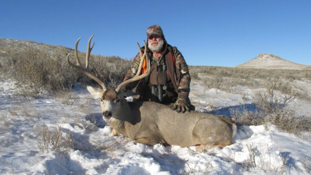 A hunter kneels next to a harvested mule deer in a snowy field.