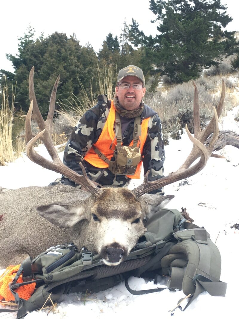A hunter poses with a harvested mule deer in a snowy, wooded landscape.