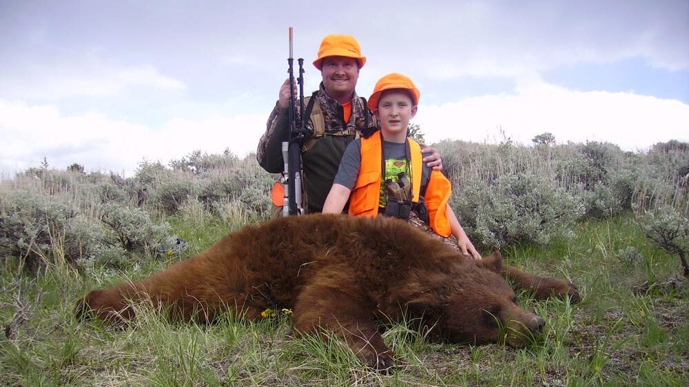 A man and a boy pose with a harvested black bear in a grassy, brushy field.