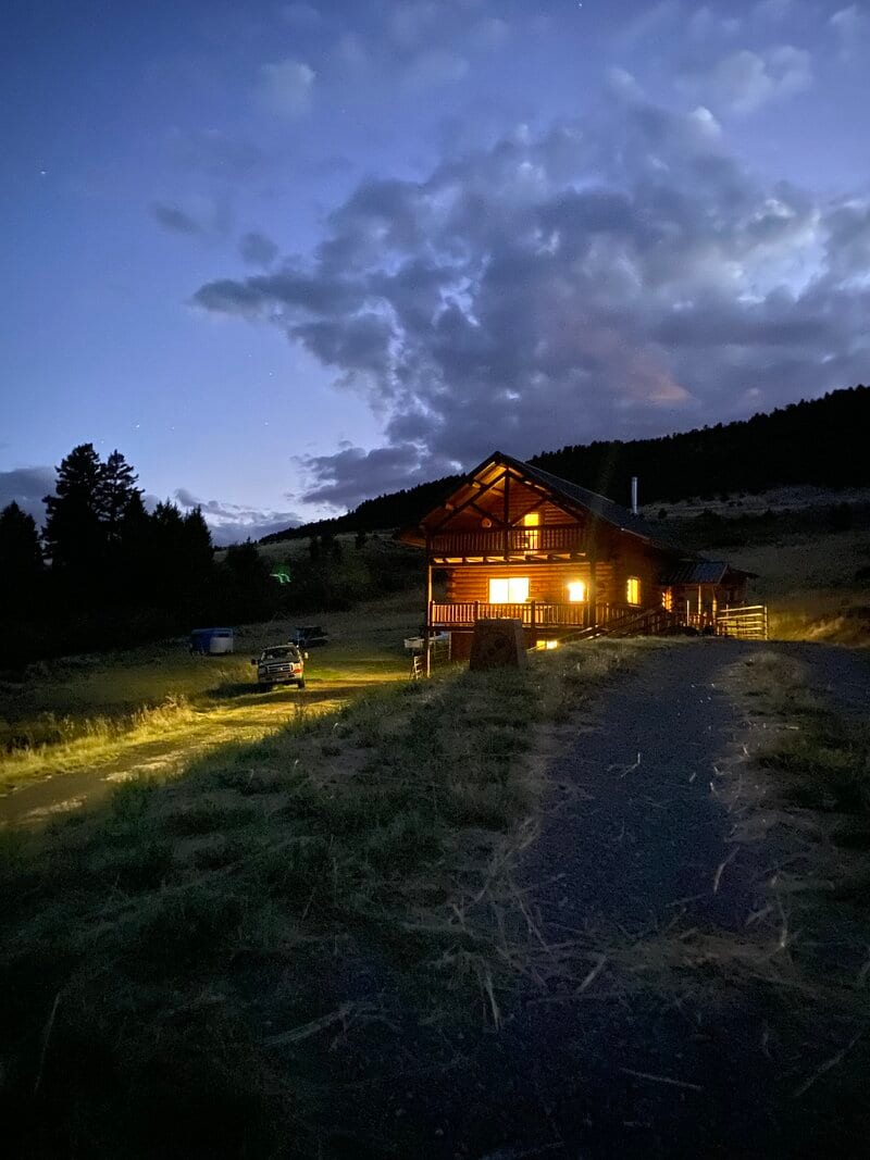 A rustic log lodge is illuminated at night under a cloudy sky, with a vehicle parked on a dirt road nearby.