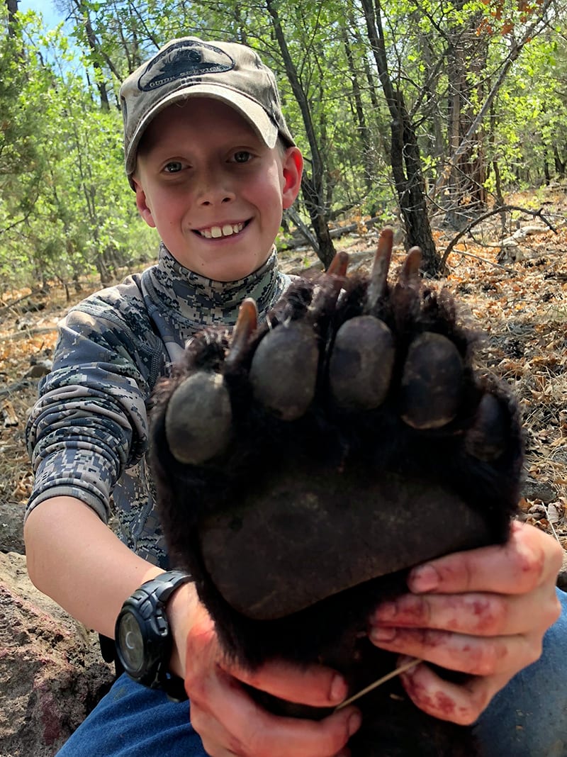 A young boy smiles while holding a black bear paw, with some blood on his hands.