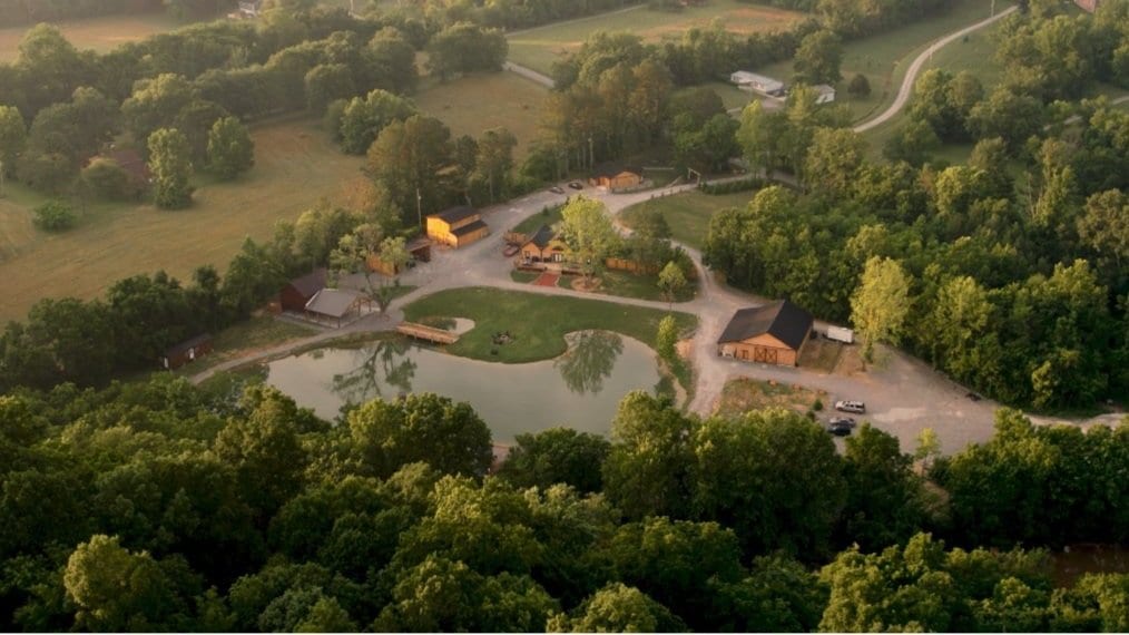 An aerial view of a property featuring several buildings, a pond, and roads surrounded by trees and fields.