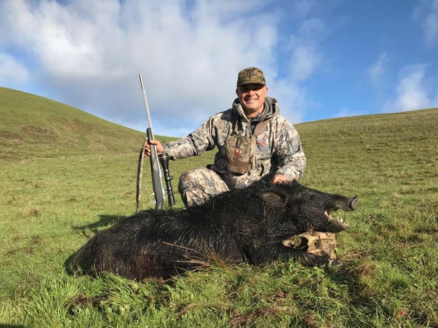 A hunter in camouflage poses with a harvested wild boar in a grassy, hilly field under a partly cloudy sky.