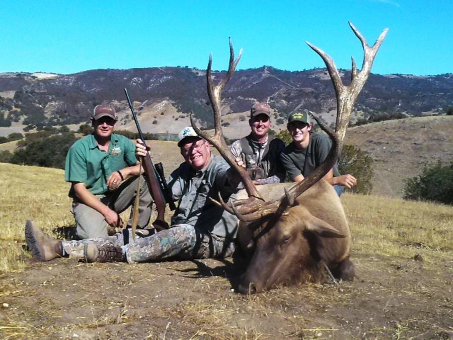 Four hunters pose with a harvested elk in a field.