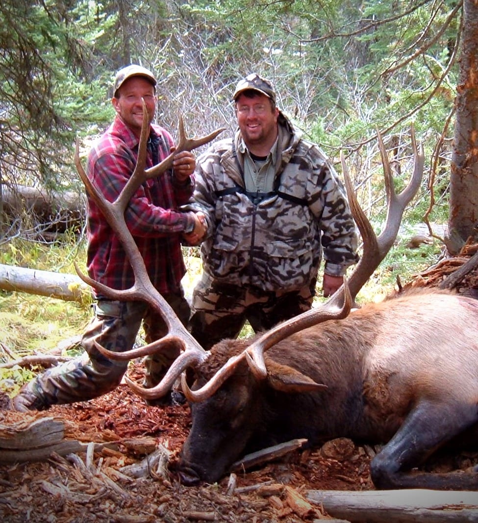 Two hunters pose with a harvested elk in a forest.