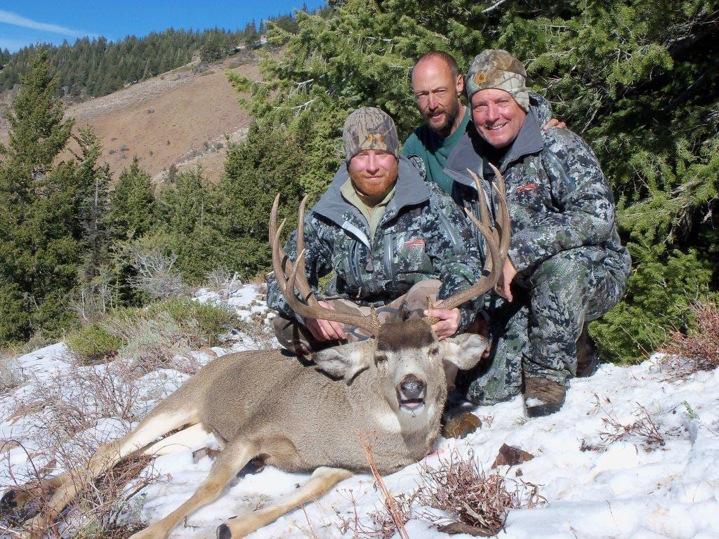 Three hunters pose with a harvested mule deer in a snowy, forested landscape.