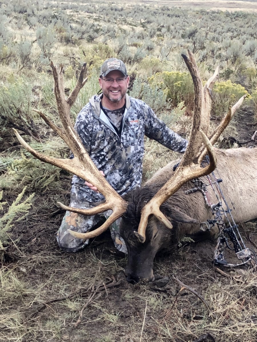 A hunter in camouflage kneels next to a harvested elk with large antlers in a field.